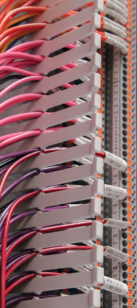 Close-up of a series of pink, red, and purple wires neatly connected to white terminal blocks in a control cabinet. The terminals are labeled with small tags, and orange details mark the contacts.