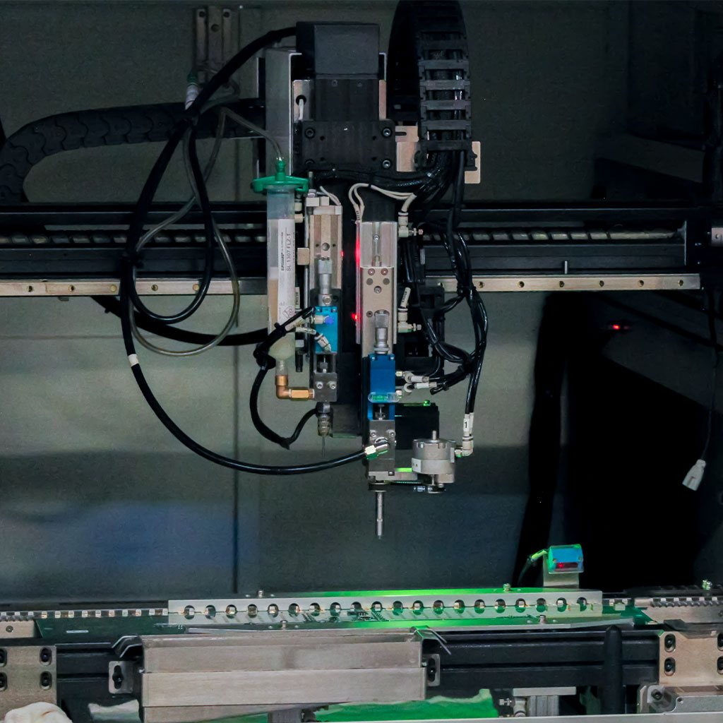 Two people wearing black work clothes and gloves are lifting two ventilated plastic cases together on a workbench in a workshop. Industrial equipment and a large fan can be seen in the background.