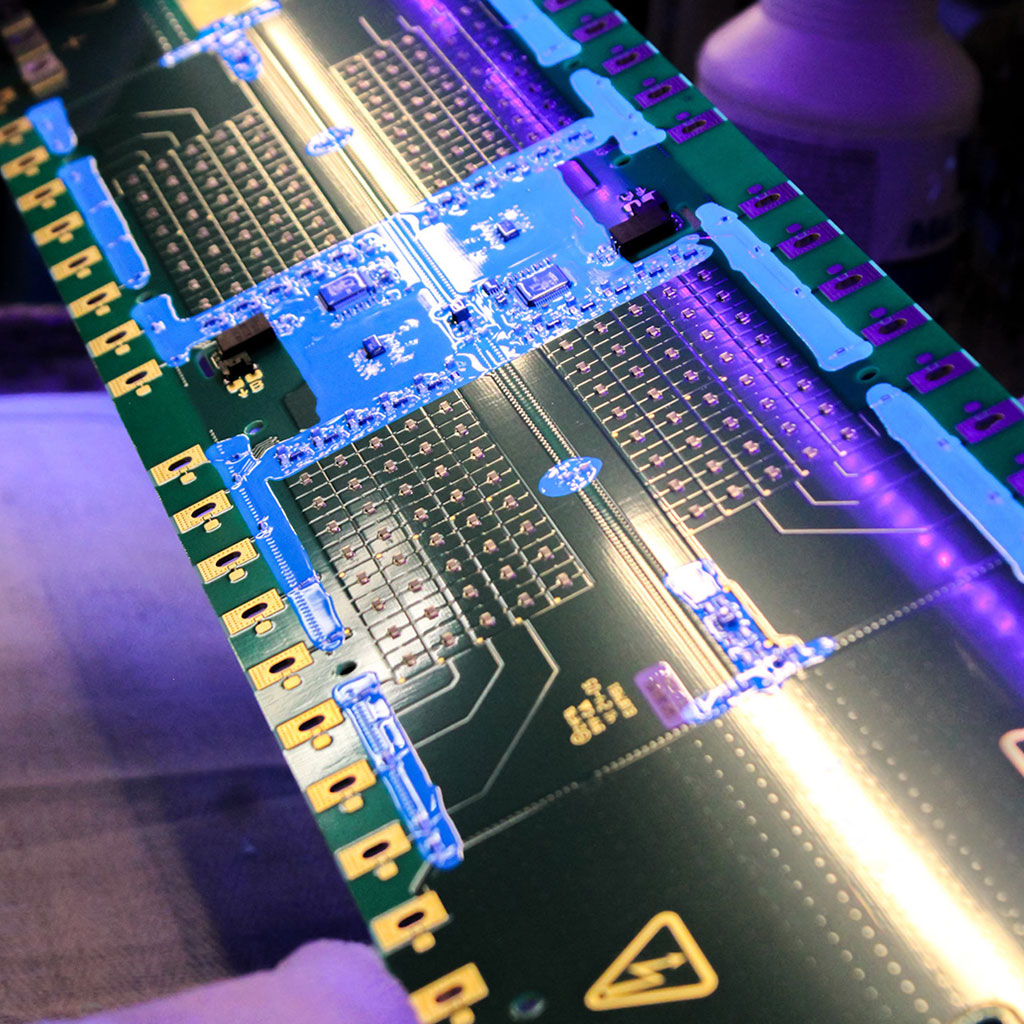 Close-up of a green circuit board with complex conductor tracks, chips, and electronic components. Blue solder and resin lines run across the board, glowing under violet UV light.